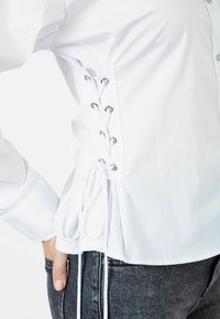 White cotton blouse featuring lace-up design with silver eyelets on the sides, and adjustable tie detail at the waist.