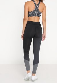 Leggings neri con un pannello di accentuazione grigio alla caviglia, abbinati a un reggiseno sportivo a disposizione racerback con motivo. Il tessuto è liscio e aderente.