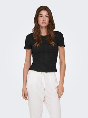 Frau mit langen braunen Haaren, die ein schwarzes, kurzärmeliges, strukturiertes Oberteil und weiße Hose mit Kordelzug trägt, stehend vor einem schlichten, hellen Hintergrund.