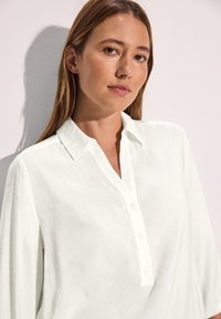 Camisa blanca con cuello, botonera, mangas largas y una tela texturizada. Diseño simple con una apariencia ligera y aireada.