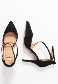 Chaussures à talons hauts en suede noir avec un bout pointu et une bride de cheville. Elles disposent d'une semelle intérieure beige et d'un talon aiguille élancé.