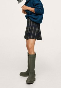 Groene rubberen kniehoge laarzen met een gestructureerd oppervlak en dikke zolen, gecombineerd met een donker geruite plooienrok en een blauwe gebreide trui.