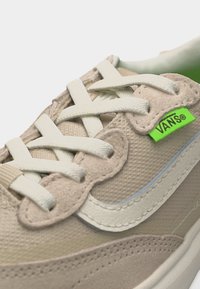 Μπεζ αθλητικό παπούτσι Vans με λευκά κορδόνια, υφές από σουέντ και δίχτυ, και πράσινη ετικέτα με το λογότυπο Vans κοντά στις τρύπες των κορδονιών.