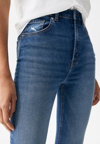High-waisted, slim-fit denim jeans in medium blue with subtle distressing, five-pocket design, and gold stitching accents.