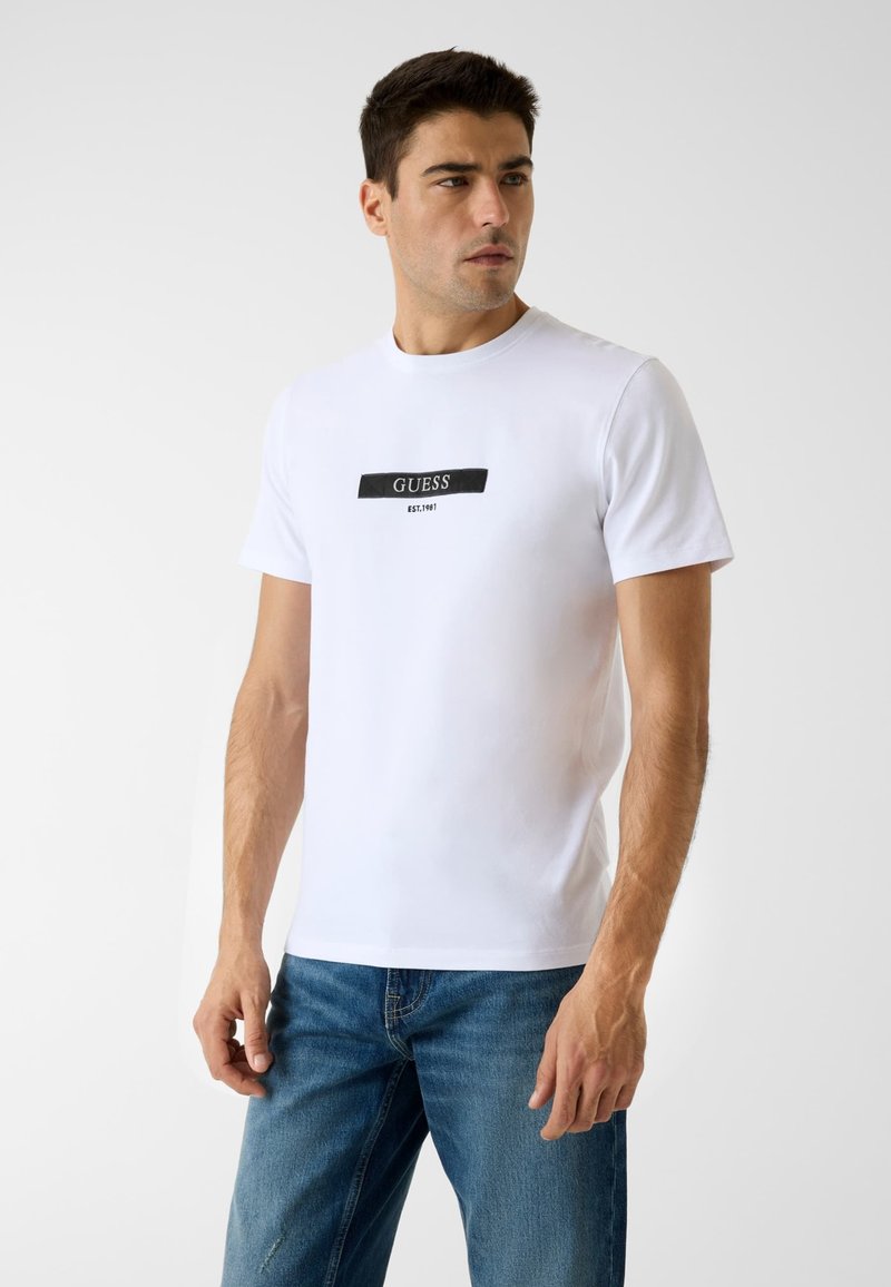 Valkoinen lyhythihainen puuvillainen t-paita, jossa musta suorakaiteen muotoinen "GUESS"-logo ja "EST. 1981" -teksti painettuna rinnan keskelle.