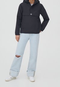 Chaqueta de sudadera con capucha negra, con un bolsillo frontal, combinada con jeans rasgados de color azul claro y zapatillas blancas. Textura suave, diseño casual.