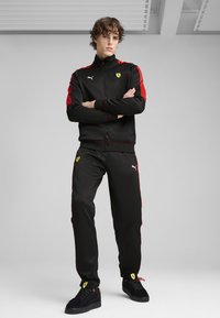 Zwart trainingspak met rode accenten, voorzien van een hoge kraag, ritsluiting en logo's. Zwarte sneakers met rode en gele details maken de look compleet.