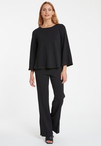 Schwarze, ausgestellte Hose mit hoher Taille und Schlitzen, kombiniert mit einem lockeren schwarzen Oberteil mit Glockenärmeln und rundem Ausschnitt aus einem weichen Stoff.