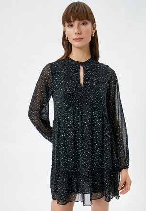 Vestido negro con lunares, mangas largas y transparentes, escote redondo con detalle de abertura y dobladillo con volantes en capas. Tejido ligero.