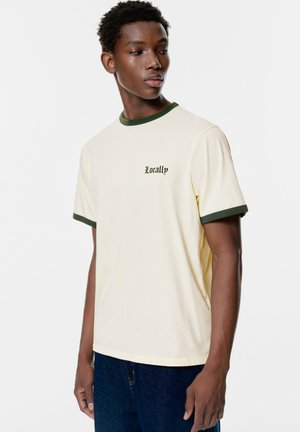 Tricou de culoare crem cu guler și accente ale manșetelor verde, având brodat "Locally" în verde închis pe piept.