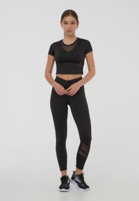 Top corto sportivo nero con dettagli in rete abbinato a leggings a vita alta con pannelli in rete e una texture liscia. Sneakers nere.