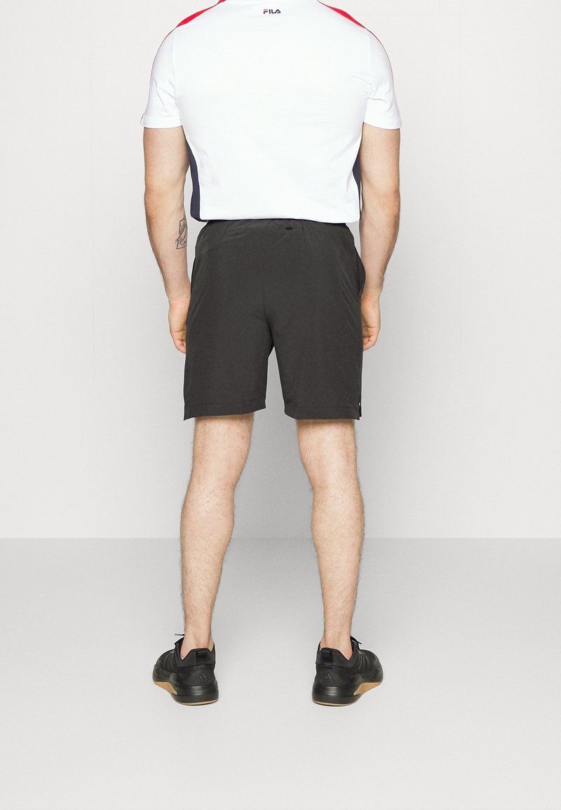 Hombre de pie frente a una pared, vestido con una camiseta de manga corta blanca y roja, pantalones cortos negros y zapatillas deportivas negras con suelas de goma.