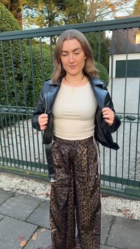 Fekete bőrkabát, bézs testhez simuló felső és barna leopárdmintás melegítőnadrág fehér oldalsávokkal. Kültéri környezet zöld kapuval.