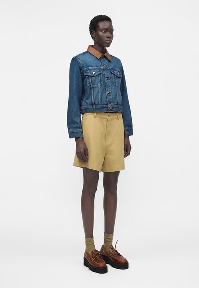Γυναίκα με κοντά μαλλιά που φοράει μπλε τζιν μπουφάν με καφέ γιακά, μπεζ σορτς μέχρι το γόνατο, καφέ κάλτσες και καφέ παπούτσια με πλατφόρμα.