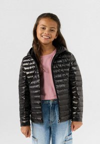 Chaqueta acolchada negra con superficie brillante y acolchada, capucha y cierre frontal. Combinada con una camisa rosa clara y pantalones de mezclilla.