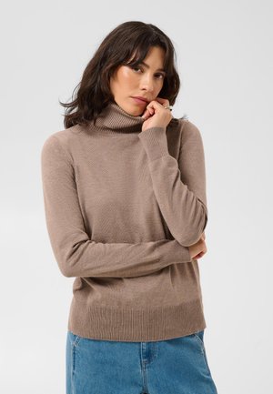 Mujer con cabello oscuro que lleva un suéter de cuello alto beige y vaqueros azules, apoyando la barbilla en la mano, con expresión pensativa.