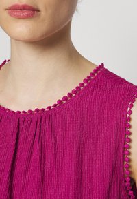 Femme portant un haut sans manches magenta texturé avec un col rond orné de petits pompons en tissu, lèvres visibles avec un rouge à lèvres rose doux.