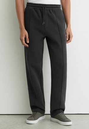 Schwarze Jogginghose mit elastischem Bund, mit seitlichen Nahtdetails, einem lockeren Schnitt, kombiniert mit grauen Sneakers vor hellem Hintergrund.