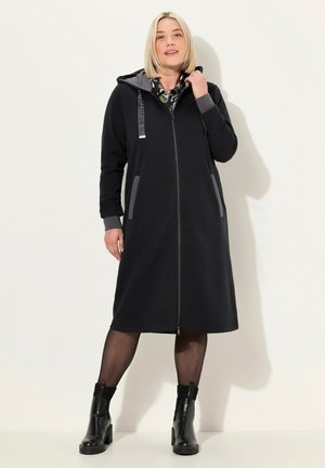 Czarna sukienka-hoodie z zamkiem, długimi rękawami, szarymi akcentami i bocznymi kieszeniami. Noszona z czarnymi botkami za kostkę i przejrzystymi legginsami.