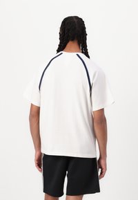 Hvid kortærmet t-shirt med marineblå detaljer på skuldrene. Blødt bomuldsstof, afslappet pasform. Båret med sorte shorts, set fra ryggen.