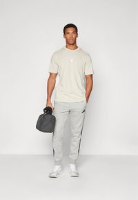 Béžové športové tričko s logom Adidas, sivé tepláky s čiernymi bočnými pruhmi a biele tenisky. Osoba drží čiernu športovú tašku.