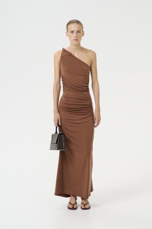 Brown one-shoulder dress made of stretchy material, featuring ruched detailing throughout and a floor-length hem. Model holds a small black handbag.