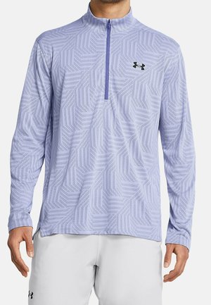 Mand iført lyse lilla langærmet quarter-zip trøje med geometrisk mønster og sort Under Armour-logo på brystet, kombineret med lysegrå shorts.