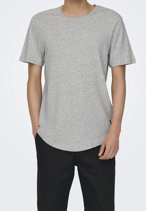 Mann trägt ein einfarbiges hellgraues kurzärmeliges T-Shirt und schwarze Hosen, steht mit einer Hand in der Tasche und der anderen entspannt.