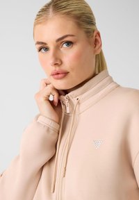 Zipperjack in zachte perzikkleurige stof met een hoge kraag, voorzien van een driehoekig strasspatroon op de borst en elastische manchetten.