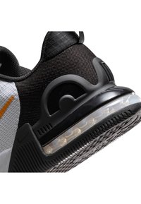 Zapatilla deportiva en blanco y negro con parte superior de malla, cuello acolchado y amortiguación de aire transparente en la suela. Presenta detalles de acento en color naranja.