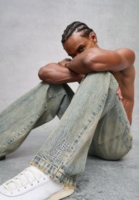 Jeans de mezclilla azul claro con un corte holgado, que presentan bordes deshilachados y un logo bordado en la parte inferior de la pierna, combinados con zapatillas blancas.