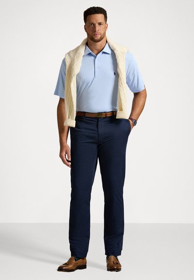 Homem com uma camisa polo azul clara, calças azul-marinho, cinto e sapatos castanhos, com um suéter em malha creme sobre os ombros, contra um fundo branco.