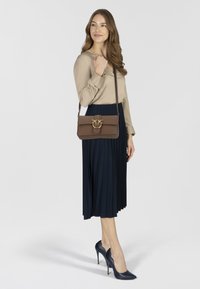 Borsa a tracolla in pelle marrone con dettaglio in metallo dorato, abbinata a una gonna plissettata blu navy e una camicetta beige. Scarpe con tacco alto blu navy.