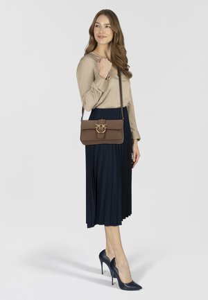 Bolso cruzado de cuero marrón con un acabado de hardware en tono dorado, combinado con una blusa beige y una falda plisada azul marino, usado con tacones altos azul marino.
