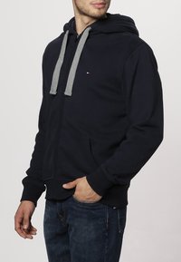 Hombre vistiendo una sudadera con cremallera azul marino y cordones grises, con jeans azul oscuro, con la mano descansando en el bolsillo frontal.