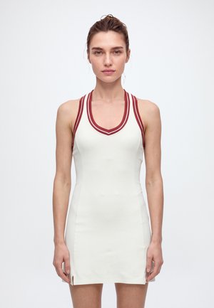 Une femme présente une robe blanche sans manches avec une bordure à rayures rouges sur un fond clair uni.