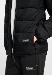 Persoon draagt een zwart gewatteerd jack en zwarte joggingbroek, beide met witte "hummel"-logo en tekst, hand in de zak van het jack.