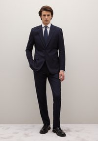 Marineblauw kostuum met krijtstreep en een getailleerde pasvorm, witte overhemd en een gestructureerde blauwe stropdas. Zwarte nette schoenen maken de formele look compleet.