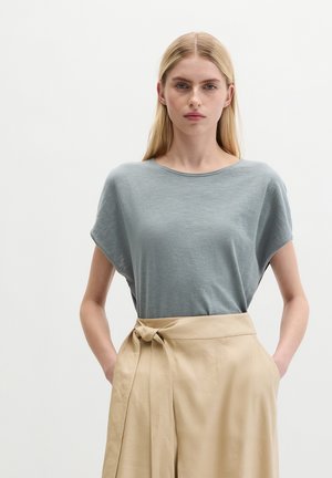 Kobieta o długich blond włosach, ubrana w luźny szary top oraz beżową spódnicę z wysokim stanem i związanym paskiem, ręce w kieszeniach, neutralne tło.