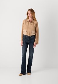 Beige langermet bluse med krage og to brystlommer, kombinert med mørkeblå utsvingte jeans og vevde beige sko på en nøytral bakgrunn.