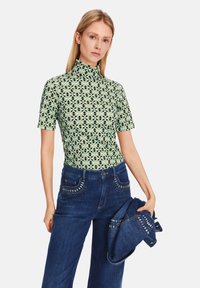 Grüner gemusterter Kurzarm-Top mit hohem Kragen, kombiniert mit dunkelblauen Jeans aus Denim, die dekorative Nieten an den Taschen haben.