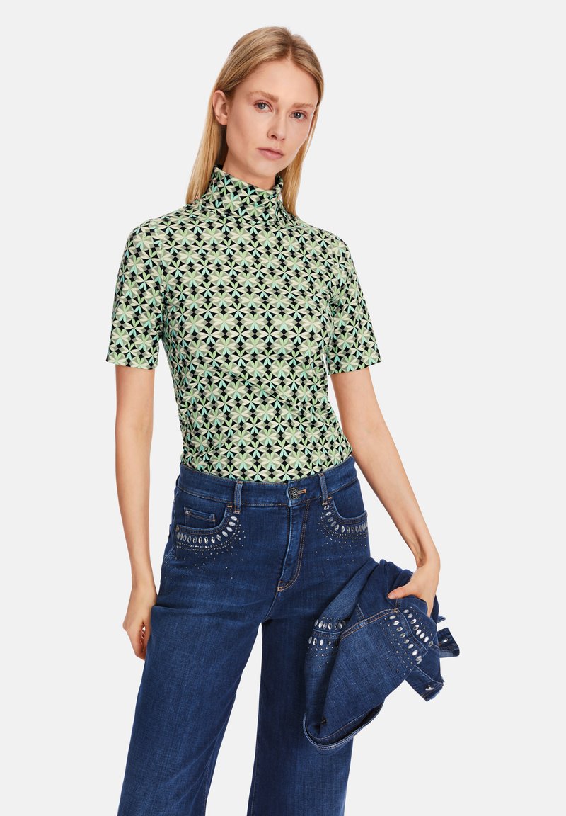 Grüner gemusterter Kurzarm-Top mit hohem Kragen, kombiniert mit dunkelblauen Jeans aus Denim, die dekorative Nieten an den Taschen haben.