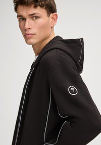 Zwarte rits-hoodie van textuurstof, met witte accentstiksels, een getailleerd ontwerp en logodetail op de linkermouw.