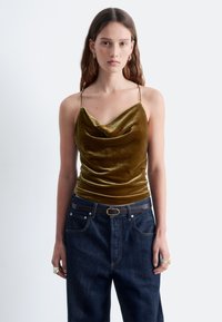 Olivgrüne Samtcami mit wasserfallartigem Ausschnitt, dünnen Trägern, kombiniert mit dunklen Jeans und einem braunen Gürtel. Besitzt einen leicht schimmernden und weichen Griff.