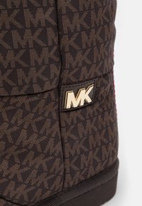 Sac en tissu marron avec un motif MK intégral, doté d'une étiquette logo MK en or, d'une base plate et d'accents rouges contrastants sur les côtés.
