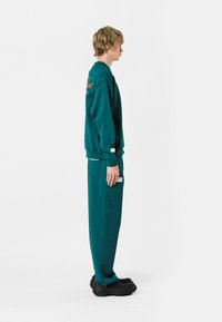 Sudadera verde azulado y pantalones a juego hechos de un tejido suave. La sudadera tiene texto bordado y los pantalones cuentan con bolsillos laterales. El calzado es negro texturizado.