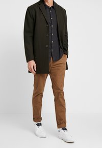 Tummanvihreä takki, jossa viisi nappia, laivastonsininen paita, jossa pienet napit, vaaleanruskeat housut ja valkoiset lenkkarit rentoon seisoma-asentoon.