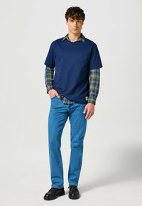 T-shirt navy sovrapposto a una camicia a quadri verde e blu. Jeans blu e scarpe nere completano l'outfit. Design semplice e informale.