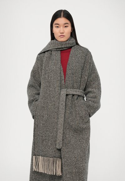 Manteau gris à motif en chevron avec une ceinture et un ourlet frangé, associé à une écharpe grise assortie. Intérieur rouge et design sans bouton.