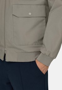 Giacca bomber grigio chiaro con cerniera frontale, polsini a costine e una grande tasca con bottone a pressione. Materiale liscio con texture minima.